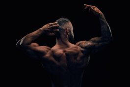 Foto von Alex Sobotta: Ein Bodybuilder mit Tattoos posiert mit dem Rücken zur Kamera, während er die Arme anspannt. Das Licht hebt eindrucksvoll seine definierten Rückenmuskeln vor dunklem Hintergrund hervor. Kraftvoll, fokussiert – Fotografie von Alex Sobotta. This is a photo of a tattooed male bodybuilder flexing his back muscles in dramatic lighting by photographer Alex Sobotta. Powerful physique and intense mood are highlighted. Alex Sobotta Fotograf