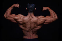 Foto von Alex Sobotta: Bodybuilding-Model Vito steht mit dem Rücken zur Kamera, Arme kraftvoll angewinkelt und Muskeln angespannt. Die gezielte Lichtführung betont jede Kontur vor dunklem Hintergrund. Ausdruck von Disziplin und Stärke in Alex Sobottas Fotografie. Vito shows his muscular back and arms in dramatic light by photographer Alex Sobotta. Fitness, bodybuilding session. Alex Sobotta Fotograf