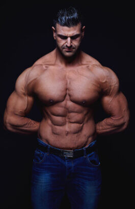 Foto von Alex Sobotta: Bodybuilding-Model Vito steht oberkörperfrei vor dunklem Hintergrund, blaue Jeans tragend. Seine definierten Bauchmuskeln und kraftvollen Arme sind akzentuiert; Blick gesenkt, Hände hinter dem Rücken. Fotografie von Alex Sobotta. A muscular male fitness model posing shirtless in jeans against a dark backdrop. Strong moody lighting highlights his physique. Alex Sobotta Fotograf