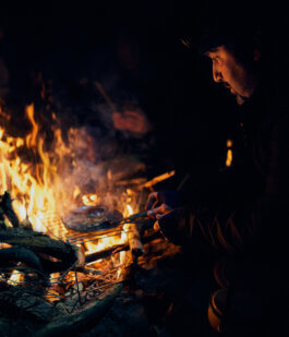 Foto von Alex Sobotta: Eine Person sitzt konzentriert am Lagerfeuer in der nächtlichen Dunkelheit, grillt ein Wagyu-Steak auf einem Gitterrost. Das warme Licht des Feuers taucht Gesicht und Hände in goldene Töne, während die Umgebung dunkel bleibt. Documentary-style photograph by Alex Sobotta showing a person tending steak over a nighttime campfire. Alex Sobotta Fotograf