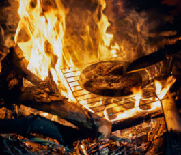 Foto von Alex Sobotta: Eine wagyu beef steak brät in einer gusseisernen Pfanne, positioniert über lodernden Holzscheiten auf einem rustikalen Metallrost. Funken und Flammen vermitteln eine authentische Lagerfeuer-Atmosphäre. Fotografie – Alex Sobotta. A rustic outdoor campfire scene with wagyu beef steak cooking over open fire; wood logs and grill add to the documentary feel. Alex Sobotta Fotograf
