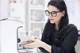 Foto von Alex Sobotta: Model Eva Maria Trummer betrachtet konzentriert eine Brille, die sie in einer modernen optischen Maschine hält; weiches Tageslicht fällt durch große Fenster auf ihren ernsten Ausdruck. Im Hintergrund technische Geräte. This is a photo of Eva Maria Trummer handling eyeglasses with precision for DAS-HANDWERK by photographer Alex Sobotta. Modern workspace, focused mood. Alex Sobotta Fotograf