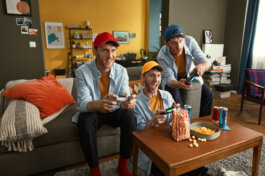 Drei Männer in passenden Outfits sitzen entspannt nebeneinander auf einem gemütlichen Sofa und spielen Videospiele. Ihr Gesichtsausdruck zeigt Konzentration und Freude, während sie zwischendurch Snacks knabbern und aus Gläsern trinken. Die warme Beleuchtung des Raumes hebt die lebhaften Farben der Einrichtung hervor; im Hintergrund sind geschmückte Regale voller Bücher zu sehen, was dem Raum eine wohnliche, einladende Atmosphäre verleiht. Die Fotografie mit dokumentarischem Charakter von Alex Sobotta („Foto von Alex Sobotta“) hält diesen Moment der Freundschaft und Geselligkeit authentisch fest: lässige Körperhaltungen, realistische Lichtführung sowie dezente Requisiten wie Controller und Snackschalen betonen das spontane Gefühl der Szene. Das Bild ist Teil einer Kampagne für Fritz!Box von Agentur: DAS KARTELL. Three friends gaming together on a couch in a colorful living room—photo by Alex Sobotta for the Fritz!Box campaign. Cozy vibes and playful energy create an inviting group scene. Alex Sobotta Fotograf