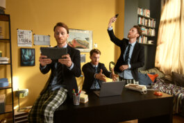 Foto von Alex Sobotta, Fotografie: Drei identische Männer – jeder trägt einen Blazer kombiniert mit karierten Pyjamahosen – befinden sich in einem hellen, modernen Homeoffice. Einer bedient konzentriert ein Tablet, der Zweite gestikuliert lebhaft am Laptop, während der Dritte mit einem leichten Lächeln ein Selfie aufnimmt. Umgeben sind sie von einer Vielzahl an Büchern und verspielter Dekoration; das Tageslicht flutet sanft durch große Fenster und betont die freundliche Arbeitsatmosphäre. Im Hintergrund sind typische Büroaccessoires zu erkennen. Produktion: Zeitsprung — Alex Sobotta fotografierte diese Szene für eine Fritz!Box Kampagne. Three men who look exactly alike pose in a cozy office for a Fritz!Box campaign by Alex Sobotta. Each interacts with different digital devices surrounded by books and personal items, creating an energetic yet homey mood. Alex Sobotta Fotograf