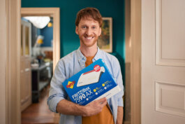 Foto von Alex Sobotta: Ein lachender Mann steht im Innenraum und hält eine blaue Fritz!BOX 7590 AX Router-Verpackung vor sich. Die Lichtstimmung ist weich und natürlich, betont die freundlichen Gesichtszüge des Mannes, dessen Körperhaltung offen und entspannt wirkt. Im Hintergrund sind petrolfarbene Wände sowie weiße Türen zu erkennen, einige Möbelstücke deuten auf einen häuslichen Kontext hin. Die Bildsprache von Alex Sobottas Fotografie fängt authentische Freude in einem modern eingerichteten Raum ein. Produktion: Zeitsprung, Agentur: DAS-KARTELL. A man stands indoors holding a blue Fritz!BOX router box and smiling at the camera. The room has teal walls and white doors; production by Zeitsprung and agency DAS-KARTELL. Alex Sobotta Fotograf