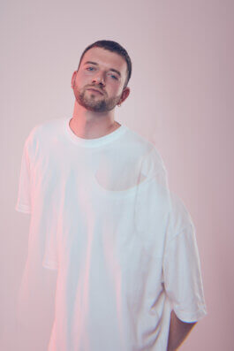 Foto von Alex Sobotta: Ein Mann mit kurzem, dunklem Haar und gepflegtem Bart, Jan-Baumeister, steht in entspannter Pose vor einem hellrosa Hintergrund. Er trägt ein lockeres weißes T-Shirt. Ein künstlerischer Doppelbelichtungseffekt dominiert die Fotografie; der Mann erscheint verschwommen und doppelt abgebildet, was dem Bild eine filmische Tiefe verleiht. Sein Blick ist nachdenklich in die Ferne gerichtet, das Licht ist weich und lässt Konturen sanft verschwimmen. Keine weiteren Requisiten oder Objekte sind im Bild sichtbar – der Fokus liegt ganz auf Ausdruck und Technik im Porträt. Der Hintergrund bleibt durchgehend gleichmäßig rosa ohne Ablenkungen. Alex Sobotta photography: A man stands against a pink backdrop with a striking double exposure effect that creates motion and emotion. Cinematic portrait with creative blur and minimalistic styling. Alex Sobotta Fotograf