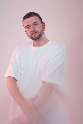 Foto von Alex Sobotta: Ein Mann, Jan-Baumeister, mit kurzem Haar und gepflegtem Bart steht vor einem sanft rosafarbenen Hintergrund. Er trägt ein lockeres, weißes T-Shirt und hält seine Arme entspannt vor dem Körper. Der Ausdruck auf seinem Gesicht ist ruhig und nachdenklich, während sein direkter Blick in die Kamera eine stille Präsenz erzeugt. Das Licht fällt weich auf ihn und hebt die klaren Konturen seines Gesichts hervor. Die Szenerie wirkt minimalistisch; keine Requisiten lenken ab – der Fokus liegt ganz auf der Person im Porträtstil dieser Fotografie von Alex Sobotta. Portrait of Jan-Baumeister by photographer Alex Sobotta with a calm and thoughtful expression against a soft pink background. Documentary-style photo showing minimalist aesthetics and relaxed posture. Alex Sobotta Fotograf