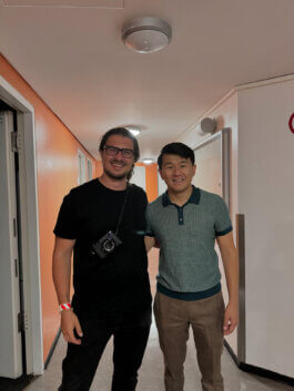 Ronny Chieng und Alex Sobotta stehen entspannt nebeneinander in einem hell erleuchteten Flur mit orange-weißen Wänden. Die Lichtstimmung ist freundlich und betont ihre lächelnden Gesichter. Einer trägt eine moderne Brille, ein schwarzes T-Shirt und hat eine Kamera um den Hals – er strahlt die Ruhe eines erfahrenen Fotografen aus; der andere trägt ein hellblaues Hemd sowie braune Stoffhosen, seine lockere Haltung unterstreicht die Dynamik des Moments. Im Hintergrund setzen dezente Schatten entlang der Wände zusätzliche Tiefe im . Requisiten sind minimal – Fokus liegt klar auf den Protagonisten und ihrem gemeinsamen Augenblick. Ronny Chieng and Alex Sobotta stand smiling together in a hallway with colorful walls. This candid portrait captures the spirit of friendship and creative work. Alex Sobotta Fotograf