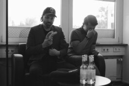 Zwei Männer sitzen in einem Innenraum auf Stühlen. Im Fokus steht ein Mann mit dunklem Bart, in stilvollem Blazer und Kappe; er hält nachdenklich eine Banane in der Hand und betrachtet sie aufmerksam. Neben ihm sitzt ein weiterer Mann, lässig gekleidet, der sein Gesicht teilweise mit der Hand verdeckt – eine unbestimmte Mischung aus Nachdenklichkeit oder Verlegenheit liegt in seiner Pose. Im Vordergrund des Bildes stehen mehrere Flaschen auf einem Tisch; das Licht ist weich und fotografisch perfekt gesetzt, wodurch Schattenspiele auf den Gesichtern entstehen. Der Hintergrund bleibt unscharf und zurückhaltend, wodurch die beiden Protagonisten im Mittelpunkt von Alex Sobottas dokumentarischer Fotografie stehen. Two men sit indoors: one gazes thoughtfully at a banana while the other covers his face. Alex Sobotta photography captures a candid moment with soft light and storytelling detail. Alex Sobotta Fotograf