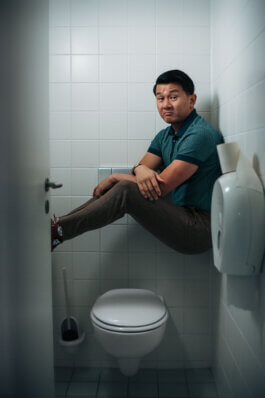 Ronny Chieng sitzt mit verschränkten Beinen und Armen, über einer weißen Toilette balancierend, eingekeilt zwischen zwei gegenüberliegenden Wänden eines kleinen Badezimmers mit weißen Fliesen. Sein Blick ist ruhig und neutral, die Mimik ernsthaft und konzentriert. Das Licht fällt von oben herein, beleuchtet Gesicht und Körper des Mannes gleichmäßig. Die Pose wirkt gespannt und ungewöhnlich akrobatisch, dabei bringt das simple Setting einen dokumentarisch-authentischen Charakter in die Aufnahme. Keine weiteren Requisiten lenken ab; der Fokus bleibt ganz auf dem Porträtierten im engen Raum. Ein typisches Beispiel für Alex Sobottas experimentelle Fotografie. A man balances above a toilet in a tiled bathroom with crossed arms and legs, holding a calm expression for an artistic portrait. This image by Alex Sobotta showcases creative documentary photography style. Alex Sobotta Fotograf