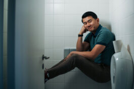 Ronny Chieng sitzt mit verschränkten Beinen und Armen, über einer weißen Toilette balancierend, eingekeilt zwischen zwei gegenüberliegenden Wänden eines kleinen Badezimmers mit weißen Fliesen. Sein Kopf ruht nachdenklich auf seiner rechten Hand, während ein leichtes Lächeln seine Lippen umspielt. Seine Beine sind entspannt ausgestreckt. Neben ihm an der Wand prangt ein auffälliger Live Nation Comedy Händetrockner. Die klare Lichtführung unterstreicht Details wie den kühlen Glanz der Fliesen und bringt eine subtil humorvolle Atmosphäre mit dokumentarischem Charakter zum Vorschein – ganz im Stil von Alex Sobotta Fotografie. Im Hintergrund bleibt die nüchterne Einfachheit des Raumes spürbar. A candid portrait of a man sitting on a toilet in a tiled restroom with a comedy-themed hand dryer visible beside him. This cinematic photo captures relaxed body language and subtle humor by Alex Sobotta photography. Alex Sobotta Fotograf