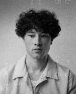 Schwarzweiß-Fotografie von Alex Sobotta: Tao, Eine junge Person mit lockigem Haar trägt eine helle Jacke und blickt leicht nach unten, der Ausdruck auf ihrem Gesicht bleibt neutral. Das Licht fällt weich über das Gesicht und betont die feinen Strukturen der Haut. Der Hintergrund ist unscharf und minimalistisch gehalten, sodass der Fokus klar auf dem Modell liegt. Die charakteristische, körnige Textur und sichtbare KODAK-Markierungen am Rand verleihen dem Bild einen dokumentarischen, zeitlosen Charakter. Foto von Alex Sobotta. A black-and-white portrait photograph by Alex Sobotta shows a young person with curly hair looking down in soft lighting and grainy Kodak film style. This image captures a timeless, vintage mood with classic photography aesthetics. Alex Sobotta Fotograf