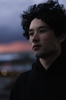 Foto von Alex Sobotta. In dieser eindrucksvollen Fotografie steht eine junge Person mit lockigem, dunklem Haar – Model: Tao – im Mittelpunkt. Ihr Blick ist nachdenklich und schweift in die Ferne, als ob sie in stiller Kontemplation versunken wäre. Das Gesicht ist sanft beleuchtet von den letzten Lichtstrahlen der Dämmerung; leichte Schatten modellieren die feinen Züge und verleihen dem Ausdruck Tiefe. Hintergrund und Umgebung verschwimmen zu einem weichen Bokeh in zarten Lila- und Rosatönen, wodurch das Motiv noch stärker hervorgehoben wird. Keine weiteren Objekte oder Requisiten lenken ab – nur Tao, das Licht der Abendstunde und eine Atmosphäre voller poetischer Ruhe prägen dieses Werk von Alex Sobotta. A cinematic portrait of a young model with curly hair at dusk by photographer Alex Sobotta. The soft purple and pink background enhances the thoughtful mood of the image. Alex Sobotta Fotograf