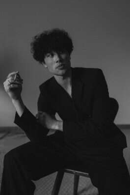 Foto von Alex Sobotta: Model Tao, mit markanten Locken, sitzt lässig in einem dunklen Anzug auf einem schlichten Stuhl. Ihr Blick ist nachdenklich zur Seite gerichtet, in der rechten Hand hält sie eine Zigarette mit eleganter Geste. Die Fotografie von Alex Sobotta ist in Schwarzweiß gehalten, weiches Licht modelliert sanft die Konturen ihres Gesichts und lässt dezente Schatten auf dem neutralen Hintergrund entstehen. Die gesamte Bildkomposition wirkt ruhig und filmisch, jedes Detail unterstreicht die zeitlose Ästhetik des Moments. Black and white documentary-style photo of model Tao by photographer Alex Sobotta, featuring classic lighting and a cinematic atmosphere. The model holds a cigarette and looks away thoughtfully while seated on a chair in a dark suit. Alex Sobotta Fotograf