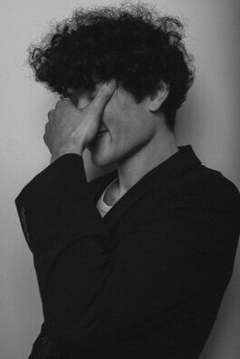 Foto von Alex Sobotta: Ein Porträt des Models Tao, das mit lockigem Haar und einem dunklen Blazer vor einem schlichten Hintergrund steht. In nachdenklicher Pose verdeckt Tao einen Teil seines Gesichts mit einer Hand, wodurch der Ausdruck von Tiefe und Introspektion verstärkt wird. Das Bild ist in Schwarzweiß gehalten, was die kontemplative Stimmung betont und den Fokus auf Licht, Schatten sowie feine Details im Gesicht legt. Die Fotografie von Alex Sobotta zeichnet sich durch dokumentarische Klarheit und cineastische Atmosphäre aus. Model Tao with curly hair in a dark blazer stands against a plain backdrop, partially covering their face in a thoughtful pose. Black and white photography by Alex Sobotta enhances the introspective mood. Alex Sobotta Fotograf