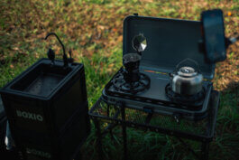 Eine mobile Outdoor-Campingküche im Stil von Alex Sobotta Fotografie: Im Zentrum steht ein robuster Gaskocher, auf dem eine klassische silberne Mokkakanne und ein emaillierter Wasserkessel dampfen. Daneben befindet sich ein schwarzer Spülbehälter mit deutlich sichtbarer BOXIO-Beschriftung – Zweckmäßigkeit trifft klares Design. Die morgendliche Szene wird von natürlichem Tageslicht durchflutet, das die glänzenden Metalloberflächen akzentuiert und einen dokumentarischen Charakter verleiht. Der Hintergrund zeigt unscharfes, sattes Grün einer Wiese – die Essenz des autarken Lebens unterwegs. Portable camp kitchen with moka pot and BOXIO sink container in fresh outdoor light; perfect for travel photography or campsite lifestyle shots. This setup captures the essence of modern nomadic living. Alex Sobotta Fotograf