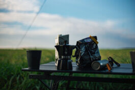 Auf einem klappbaren Campingtisch im Freien liegen eine schwarze Bialetti Moka-Kanne, eine schlichte Tasse, ein brauner Kaffeebeutel und eine kompakte Handmühle nebeneinander ausgerichtet. Die Morgensonne streift die glänzenden Metallflächen und hebt das matte Schwarz der Kanne hervor. Neben den schlicht gehaltenen Objekten ist kein Mensch sichtbar; der Hintergrund zeigt satten grünen Rasen vor weitem blauen Himmel, eingefangen mit dokumentarischer Klarheit von Alex Sobotta – Fotografie. Die Komposition vermittelt Ruhe, Authentizität und einen Hauch Freiheit. This photo by Alex Sobotta shows a moka pot and coffee gear arranged on a camping table outdoors. The scene feels relaxed and cinematic in natural morning light. Alex Sobotta Fotograf