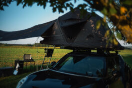 Ein schwarzer Mini Clubman steht markant im hohen Gras eines offenen Feldes, der Blickwinkel eingefangen im dokumentarischen Stil von Alex Sobotta Fotografie. Auf dem Dach thront ein aufgeklapptes Dachzelt von Roof Space mit ausladender Markise, unter der dezente Campingausrüstung platziert ist: ein faltbarer Tisch, ein einzelner Stuhl und einige Reiseutensilien deuten auf einen minimalistischen Lebensstil und die Freude am Unterwegssein. Die Morgensonne taucht das Fahrzeug in sanftes Licht, wirft Schatten ins satte Grün des Rasens. Im Hintergrund zeichnen sich vereinzelt hohe Bäume gegen den wolkenlosen blauen Himmel ab; die Szene strahlt Ruhe und Abenteuerlust zugleich aus. A black Mini Clubman with a rooftop tent and camping setup stands in a tranquil grassy field under blue skies. Documentary photography by Alex Sobotta captures the serenity and spirit of van life. Alex Sobotta Fotograf