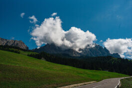 Eine kurvige Straße schlängelt sich durch eine grüne, grasbewachsene Hügellandschaft, flankiert von einem dichten Kiefernwald. Im Hintergrund ragen majestätische Berge empor, deren Spitzen teilweise von großen, flauschigen weißen Wolken verdeckt werden. Über der Szenerie erstreckt sich ein strahlend blauer Himmel, dessen Licht das satte Grün und die Textur der Landschaft hervorhebt – ein Moment eingefangen im Stil von Alex Sobotta Fotografie. Keine Menschen oder Fahrzeuge sind sichtbar; die Szene wirkt ruhig und unberührt. A winding road leads through lush hills with pine forests and mountains beneath dramatic clouds in the background. Captured in the style of landscape photography by Alex Sobotta. Alex Sobotta Fotograf