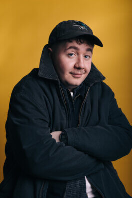 Porträtfotografie eines Mannes in dunkler Jacke und Baseballcap, der mit verschränkten Armen und einem dezenten, verschmitzten Lächeln wie Berliner Comedians vor einem schlichten gelben Hintergrund sitzt. Das Licht ist weich und betont seine entspannte Pose, während der minimalistische Hintergrund die Person in den Mittelpunkt rückt. Kein sichtbares Requisit außer seiner Kleidung. Dokumentarischer Blick auf Ausdruck und Persönlichkeit im urbanen Stil eines Berlin-Fotografen. A comedian sits with arms crossed and a slight smile in front of a yellow background. Urban portrait photography with relaxed expression and cinematic mood. Alex Sobotta Fotograf