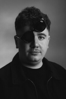 Porträt eines Mannes mit kurzen, lockigen Haaren; er trägt eine Sonnenbrille, deren linkes Glas über dem linken Auge sitzt und das rechte Glas erhöht über dem rechten Auge schwebt. Sein Blick ist neutral, fast nachdenklich, der Mund entspannt. Die Lichtführung ist sanft und legt feine Schatten auf das Gesicht sowie die dunkle Jacke des Mannes. Der Hintergrund bleibt schlicht und lenkt den Fokus ganz auf den Protagonisten – eine typische Fotografie eines Berliner Straßenfotografen, die authentische Individualität einfängt. A man poses for a portrait wearing sunglasses in an unusual way and a dark jacket. The plain background keeps the focus on his face and unique expression. Alex Sobotta Fotograf