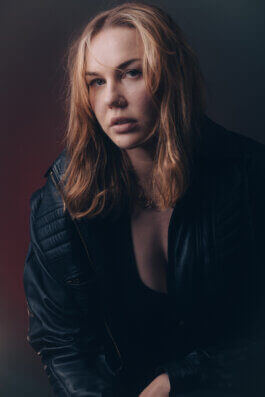 Porträtfotografie einer Person mit schulterlangem, blondem Haar und Nasenring; sie trägt eine schwarze Lederjacke und blickt mit intensiver Ernsthaftigkeit direkt in die Kamera. Das Licht ist weich und dezent, modelliert sanfte Schatten auf Gesicht und Kleidung, während ein dunkler Hintergrund den Fokus auf das ausdrucksstarke Motiv lenkt. Die Szene erinnert an klassische Berliner Streetstyle-Aufnahmen – urban, roh, filmisch. A striking blonde person with a nose ring poses in a black leather jacket against a moody background. Berlin photographer style: bold expression and cinematic lighting evoke an urban documentary feel. Alex Sobotta Fotograf