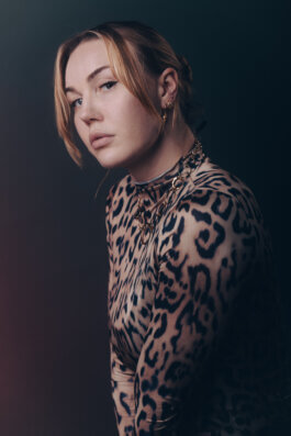 Foto von Alex Sobotta: Blondhaariges Model Clarissa mit heller Haut trägt ein Leopardenmuster-Top, große goldene Ohrringe und eine auffällige Goldkette. Ihr ernster Blick trifft die Kamera im sanften Studiolicht vor dunklem, geheimnisvollem Hintergrund. Fotografie Alex Sobotta. Clarissa poses in a moody studio portrait with gold jewelry and leopard print. Intense look and dramatic lighting create a cinematic effect. Alex Sobotta Fotograf