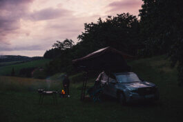 Foto von Alex Sobotta: Eine Person steht nachdenklich am Lagerfeuer neben einem 2025 Subaru Forester mit Dachzelt auf einer grünen Wiese bei Dämmerung. Campingstühle, Grill und Ausrüstung neben dem Auto, sanfte Hügel und Bäume im Hintergrund, wolkiger Himmel. Atmospheric camping photography by Alex Sobotta shows outdoor adventure and modern SUV lifestyle. Alex Sobotta Fotograf