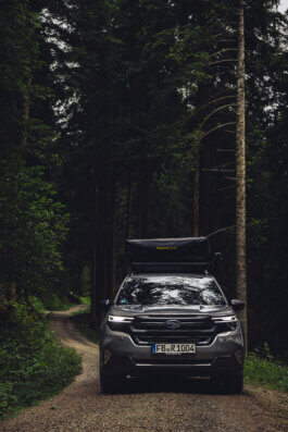 Foto von Alex Sobotta: Eine silberne 2025 Subaru Forester mit Dachzelt steht auf einem schmalen Schotterweg, umgeben von hohen, dichten Bäumen in einem ruhigen Wald. Das Auto blickt frontal zur Kamera, das Licht ist kühl und diffus. Dokumentarische Fotografie von Alex Sobotta. A silver 2025 Subaru Forester with a roof tent is parked on a narrow gravel road in a dense forest. The car faces the camera in soft, cool light. Alex Sobotta Fotograf