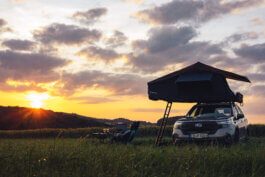 Foto von Alex Sobotta: Ein silberner Subaru Forester mit Dachzelt steht auf einer grünen Wiese im goldenen Sonnenuntergang. Zwei Campingstühle und ein Tisch mit Getränken bilden eine einladende Szenerie, eingerahmt von dramatischem Himmel und sanften Hügeln im Hintergrund. Stimmungsvolle Dokumentarfotografie von Alex Sobotta, das warme Licht betont die Ruhe des Moments. A Subaru Forester with a roof-top tent in scenic light, surrounded by camping gear and nature at sunset. This image captures outdoor adventure and tranquility. Alex Sobotta Fotograf