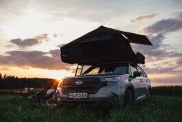 Foto von Alex Sobotta: Eine silberne Subaru Forester mit Dachzelt steht im abendlichen Licht auf einer grasbewachsenen Wiese. Zwei Campingstühle und ein Tisch mit Getränken arrangiert, umrahmt von Bäumen und dramatischer Himmel; Fotografie Alex Sobotta. A silver Subaru Forester with a rooftop tent, camping chairs, and a table in a grassy field at sunset. This cinematic car camping scene is perfect for outdoor lovers and adventure photographers. Alex Sobotta Fotograf