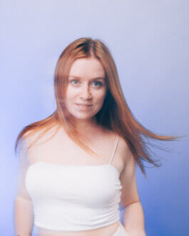 Porträt einer Frau mit langen roten Haaren, weißem Spaghettitop, vor hellblauem Hintergrund. Ihr leicht bewegtes Haar und ihr sanfter Blick fangen das Licht ein. Fotografie von Alex Sobotta. Photo by Alex Sobotta, cinematic feel. Alex Sobotta Fotograf