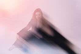 Eine Frau in dunkler Kleidung sitzt am Boden, sanftes, verträumtes Licht in Rosa legt sich wie ein Schleier über sie. Ihr Gesicht ist nur teilweise sichtbar. Foto von Alex Sobotta, künstlerische Fotografie. Woman sitting on floor with dreamy pink light by Alex Sobotta. Alex Sobotta Fotograf