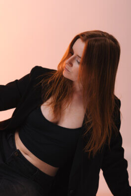 Foto von Alex Sobotta: Eine Frau mit langen roten Haaren, schwarzem Blazer und schwarzem Top sitzt vor sanft rosa Hintergrund. Ihr Blick ist zur Seite gerichtet, nachdenklich, das Licht weich. Fotografie von Alex Sobotta. A red-haired woman looks thoughtful on a pink backdrop. Alex Sobotta Fotograf