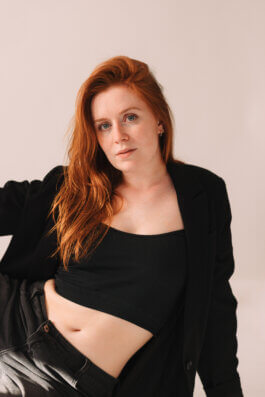 Porträt einer Frau mit langen roten Haaren, schwarzem Crop-Top, Hose und Blazer vor neutralem, hellem Hintergrund. Direkter Blick in die Kamera, neutrales Gesicht. Stimmungsvolle Fotografie von Alex Sobotta. Modern woman portrait. Simple backdrop. Alex Sobotta Fotograf
