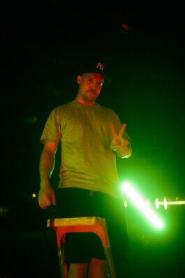 Aron steht auf einer Leiter im Velodrom, trägt graues T-Shirt, schwarze Shorts, schwarze Yankees-Cap. Er zeigt ein Peace-Zeichen, hinter ihm leuchtet grelles grünes Licht im Dunkeln. Fotografie von Alex Sobotta. Cool night portrait, peace gesture. Alex Sobotta Fotograf