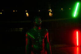 Eine nackter Aron mit glänzender Haut steht im Velodrom, dramatisch von grünen und roten Neonlichtern beleuchtet. Spiegelnde Reflexionen tanzen hinter ihr, der Hintergrund verschwimmt im Dunkel. Foto von Alex Sobotta, Fotografie. A moody portrait in neon light by Alex Sobotta. Alex Sobotta Fotograf