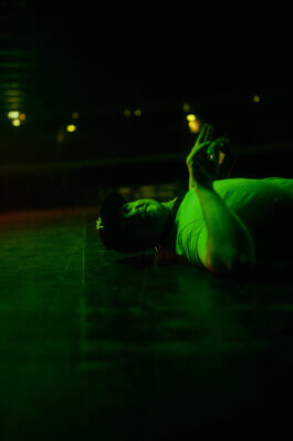 Im dokumentarischen Stil: Marco Justus Schöler mit Kappe liegt auf dem dunklen Velodrom-Boden im grünen Licht, blickt nach oben und hält einen Gegenstand. Unscharfe Lichter im Hintergrund. Fotografie, Foto von Alex Sobotta. Night, green lights, urban velodrome scene. Alex Sobotta Fotograf