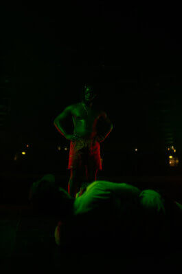 Ein muskulöser Mann in roten Shorts steht selbstbewusst mit Händen in der Hüfte im Velodrom, dramatisch von rotem und grünem Licht beleuchtet. Im Hintergrund liegt Aron am Boden im Halbdunkel. Fotografie von Alex Sobotta. Two men, dramatic lighting. Alex Sobotta Fotograf