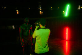 Ein fokussierter, oberkörperfreier Mann steht im Velodrom unter grüner Beleuchtung, während Marco Schöler ihn mit einer Kamera filmt. Hinter ihm leuchten vertikal grüne und rote Neonröhren. Foto von Alex Sobotta, Fotografie. Green light, cinematic portrait. Alex Sobotta Fotograf