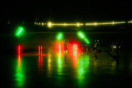 Mehrere Menschen stehen im dunklen Innenraum des Velodroms, dramatisch beleuchtet von grünen und roten Neonröhren, deren Licht sich auf dem glänzenden Boden spiegelt. Im Hintergrund leuchtet eine gelbe Kontur. Fotografie von Alex Sobotta. People in a dim venue with neon lights and reflections. Alex Sobotta Fotograf