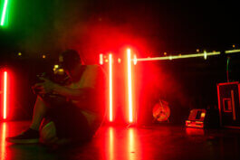 Cineastische Fotografie von Alex Sobotta: Marco Schöler mit Cap sitzt ausdruckslos auf dem Boden des schummrig beleuchteten Velodroms. Rotes und grünes Neonlicht, Bühnen-Equipment im Hintergrund. Dokumentarisch festgehalten. Moody neon-lit stage scene. Dramatic portrait of a person sitting on the floor with props. Alex Sobotta Fotograf