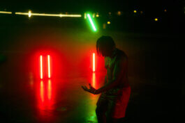 Ein oberkörperfreier Mensch steht in einem dunklen Raum, dramatisch inszeniert durch kräftige, vertikale Rot- und Grünlichter im Hintergrund. Spiegelungen tanzen am Boden. Reportageartige Fotografie von Alex Sobotta. Striking colored lights create atmosphere; Alex Sobotta photo. Alex Sobotta Fotograf