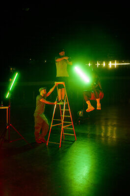 Zwei Personen richten grüne Lichter auf einer Leiter im Velodrom aus, Aron springt – unter intensivem Grünlicht in dunkler Halle sitzt. Fotografie von Alex Sobotta. Alex Sobotta Fotograf