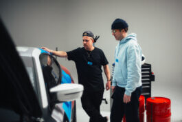 Fotografie von Alex Sobotta: Marco und Dex stehen wie Filmfiguren neben einem Auto im grellen Licht einer Werkstatt. Dex lehnt lässig am Wagen, Marco betrachtet ihn ernst. Im Hintergrund rote Fässer, Werkzeugkiste. Intense moment, industrial vibe. Alex Sobotta Fotograf