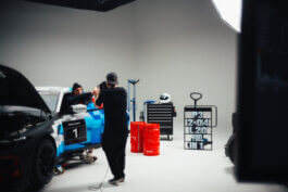 Im Studio parkt ein Rennwagen mit der Nummer 1, flankiert von Marco und Hagen. Umgeben von Werkzeugwagen, roten Fässern, Helm und Boxentafel. Atmosphäre wie bei einem Behind-the-Scenes-Fotoshooting. Fotografie von Alex Sobotta. Racing car studio photo by Alex Sobotta. Alex Sobotta Fotograf