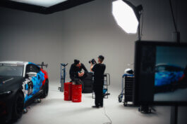 Fotografie von Alex Sobotta: Haagen sitzt entspannt mit ernstem Blick auf roten Fässern, ein Rennwagen glänzt im Hintergrund. Marco hält die Kamera, Lichttechnik und Monitore schaffen dokumentarisches Studio-Feeling. Photographic portrait, racecar studio atmosphere. Alex Sobotta Fotograf