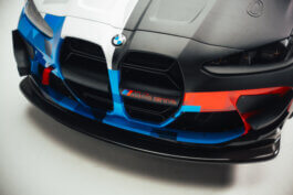 Cineastisches Close-up des BMW M4 GT4-Frontbumpers: markante Nieren, dynamische blaue und rote Rennstreifen, das M4 GT4-Emblem in Szene gesetzt. Foto von Alex Sobotta. Photographie im Studio-Licht. BMW race car details. Striking front grille shot. Alex Sobotta Fotograf