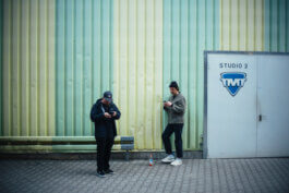 Fotografie von Alex Sobotta: Marco und Hagen, konzentriert auf ihre Smartphones, draußen vor einer gelb-grün gestreiften Wand neben der Tür „Studio 2“. Einer lehnt lässig an der Wand, zwischen ihnen eine Flasche am Boden. Candid documentary street moment. Relaxed urban scene. Alex Sobotta Fotograf