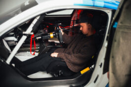 Fotografie von Alex Sobotta: Marco sitzt konzentriert im cockpitartigen Innenraum eines BMW-Rennautos, gehüllt in dunkle Kleidung und mit Kappe. Seine Hand greift nach den Schaltern am freiliegenden Armaturenbrett, Lenkrad im Vordergrund. Photo by Alex Sobotta. A racing driver checks controls inside his BMW cockpit. Alex Sobotta Fotograf